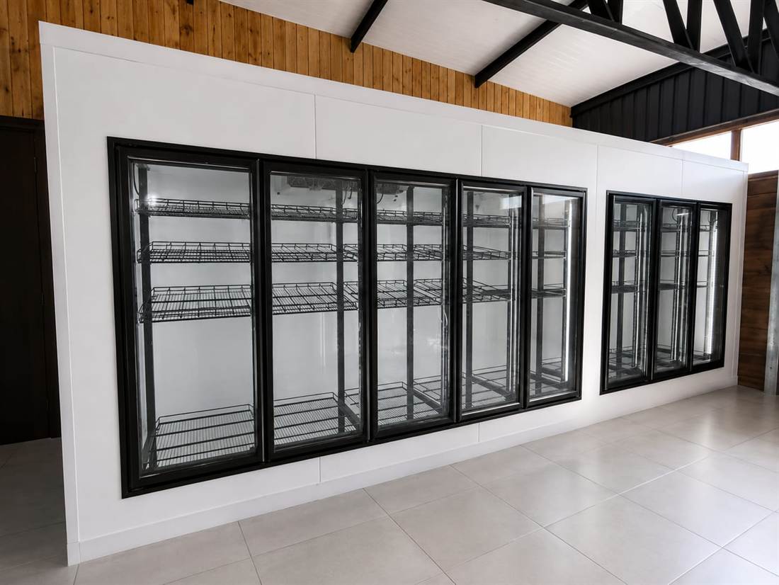 Walk-In Cooler con ocho puertas de vidrio de marco negro, estantería de alambre e iluminación LED, integrado a la pared del salón.